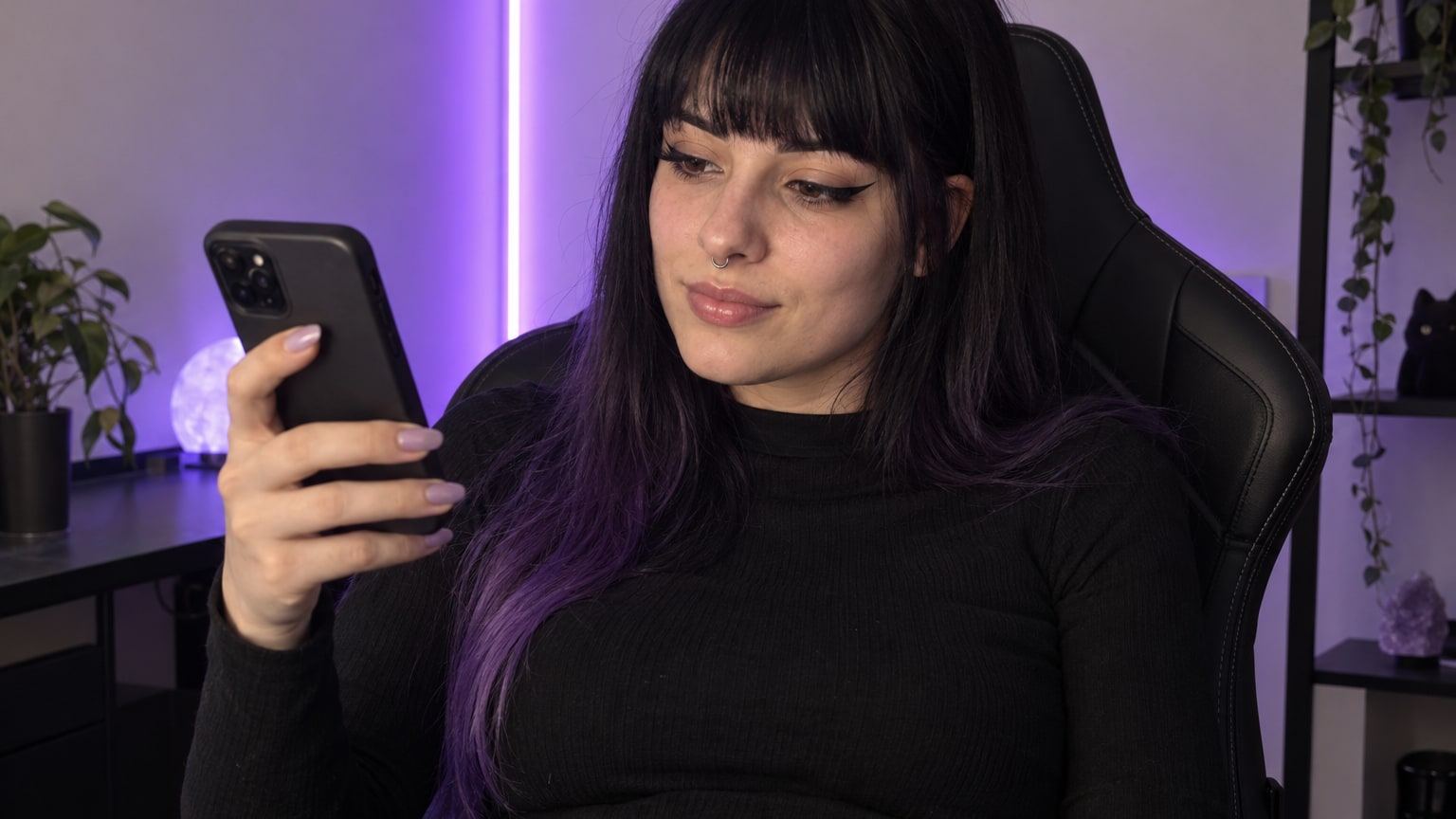 Woman with black and violet hair holds her phone in a gaming chair under purple light.