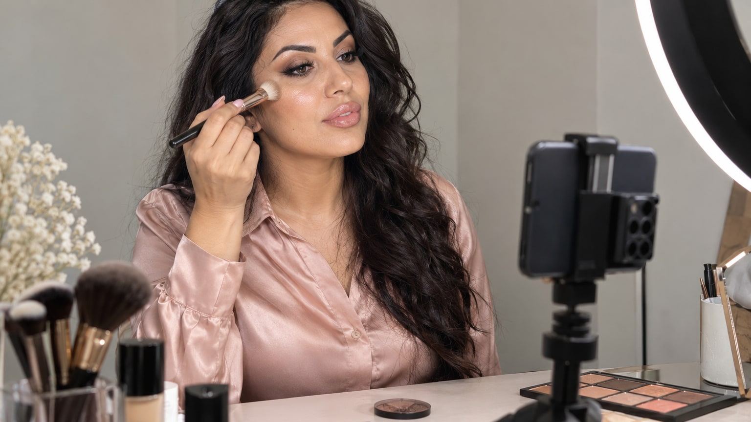Woman with long dark waves applies highlighter at a vanity lit by a ring light.