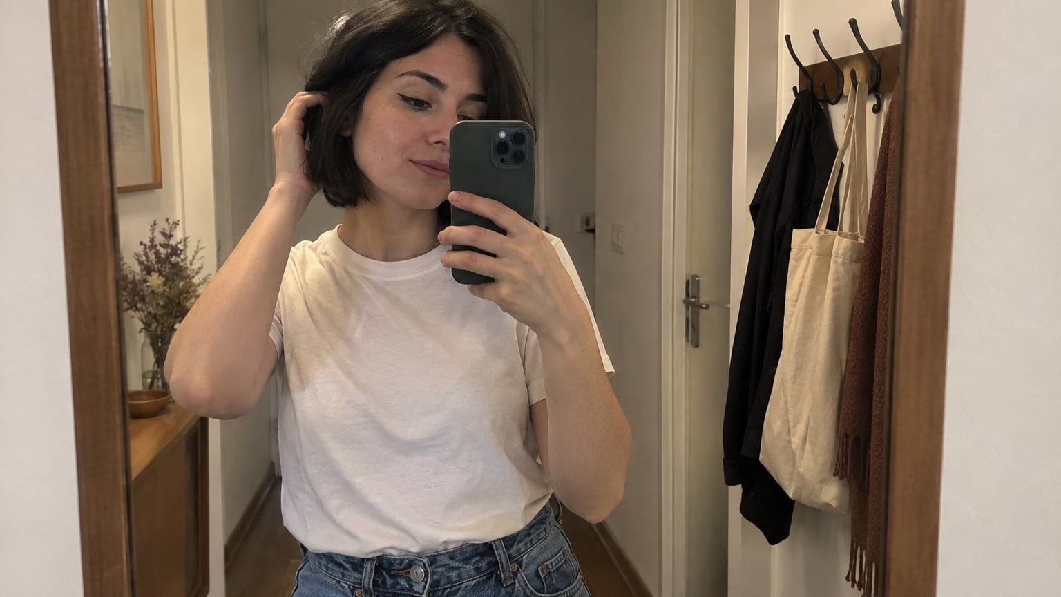 Woman with a dark bob takes a mirror selfie in a white t-shirt and jeans.