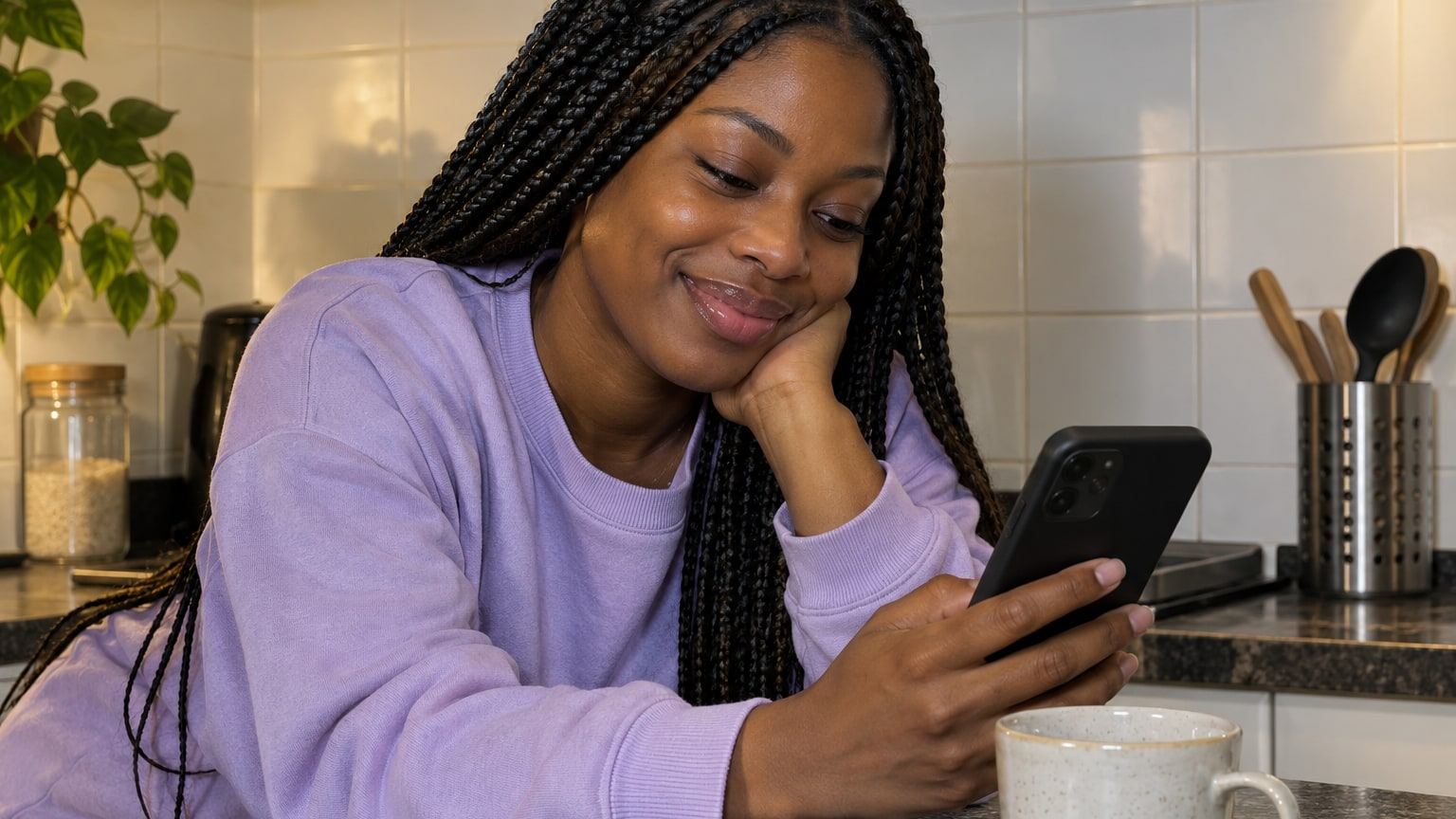 Woman with long braids smiles at her phone while leaning on a kitchen counter.