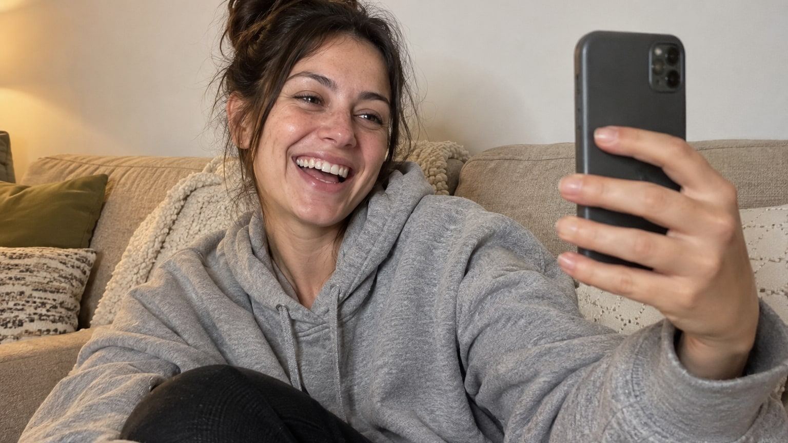 Laughing woman in a grey hoodie takes a selfie on a beige sofa.